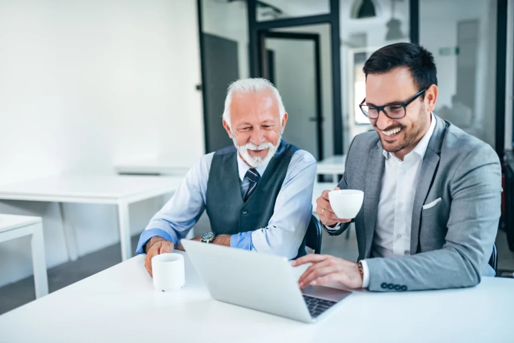 Dois profissionais sorrindo enquanto trabalham em um laptop e tomam café, simbolizando um ambiente de trabalho positivo e bem-estar financeiro.