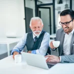 Dois profissionais sorrindo enquanto trabalham em um laptop e tomam café, simbolizando um ambiente de trabalho positivo e bem-estar financeiro.