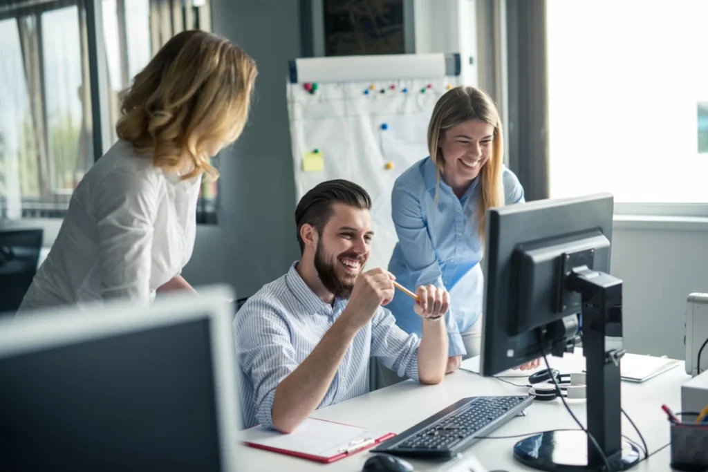 Equipe de trabalho reunida em frente a um computador, sorrindo e discutindo estratégias, representando inovação e aprendizado com IA.