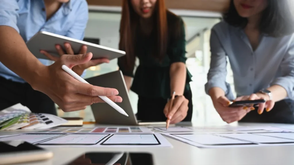 Equipe colaborando em um projeto, usando tablets, smartphones e anotações, representando a importância do trabalho em equipe, mesmo no trabalho remoto.