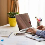 Profissional trabalhando em um laptop em um ambiente organizado, com materiais de estudo e gráficos financeiros, representando aprendizado digital.