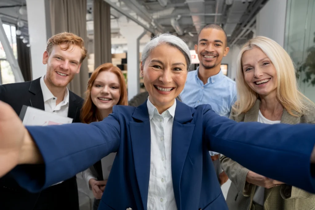 Equipe multigeracional sorrindo para uma selfie no ambiente de trabalho.