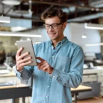 Homem sorridente usando um tablet em um ambiente de trabalho moderno, simbolizando aprendizado digital e treinamento de produto.