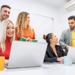Grupo de pessoas reunidas em sala de reunião com laptops, cadernos e canecas sobre a mesa, colaborando em atividades de treinamento corporativo.