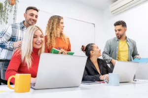 Grupo de pessoas reunidas em sala de reunião com laptops, cadernos e canecas sobre a mesa, colaborando em atividades de treinamento corporativo.