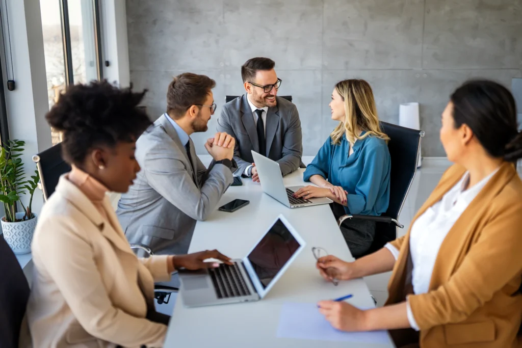 Grupo de profissionais reunidos em sala de reunião moderna, utilizando laptops e discutindo estratégias de trabalho.