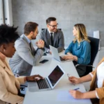 Grupo de profissionais reunidos em sala de reunião moderna, utilizando laptops e discutindo estratégias de trabalho.