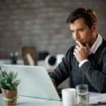 Pessoa trabalhando em um laptop sobre uma mesa com documentos, copo de água e planta decorativa, em ambiente corporativo.