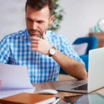 Pessoa analisando documentos enquanto trabalha em um laptop sobre uma mesa com livros e materiais de escritório.