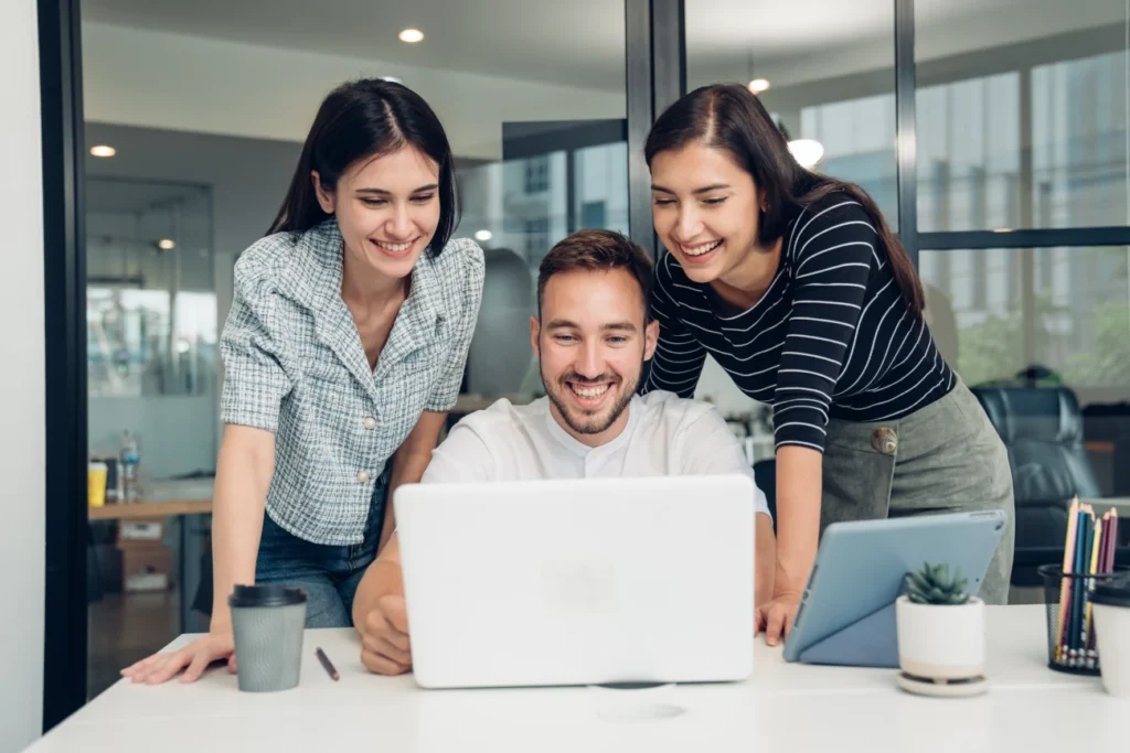 Três pessoas reunidas em frente a um laptop em ambiente corporativo, com um tablet e uma xícara sobre a mesa, simbolizando colaboração em projetos digitais.