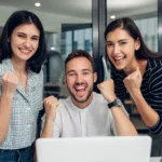 Três pessoas reunidas em frente a um laptop em ambiente corporativo, comemorando resultados ou progresso em projeto digital.