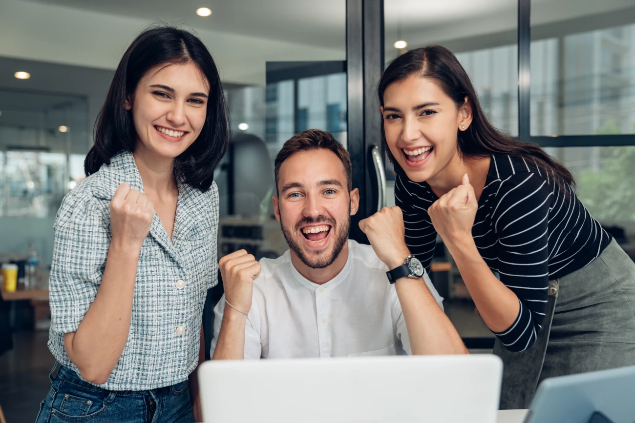 Três pessoas reunidas em frente a um laptop em ambiente corporativo, comemorando resultados ou progresso em projeto digital.