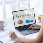 Profissional analisando gráficos de desempenho e métricas de treinamento em um laptop, com relatórios impressos sobre a mesa.