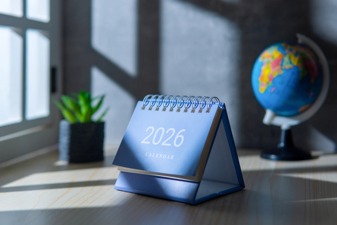 Calendário de mesa azul com o ano 2026 em destaque, posicionado sobre uma mesa próxima a um globo terrestre e uma planta decorativa.