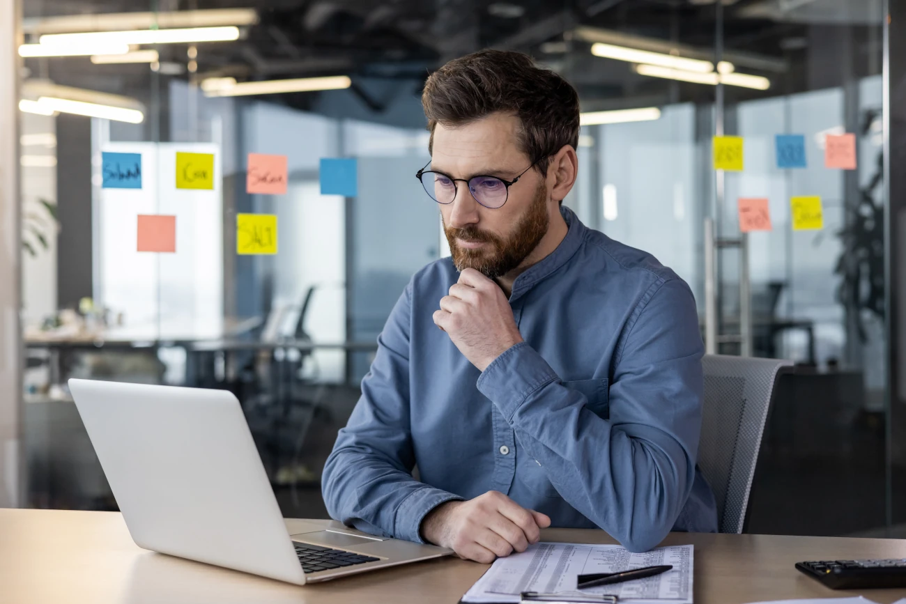 Profissional sentado à mesa de trabalho analisando informações no laptop, em um escritório com post-its coloridos no vidro ao fundo.
