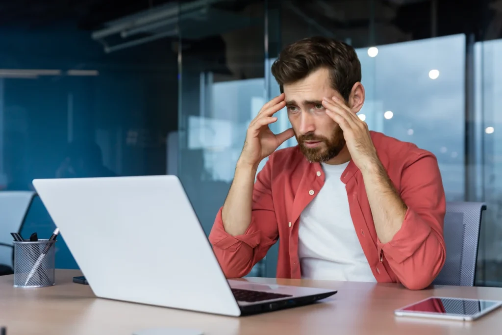 Pessoa sentada à mesa utilizando um laptop em ambiente corporativo, com as mãos apoiadas na cabeça enquanto analisa informações na tela.