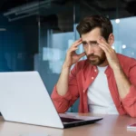 Pessoa sentada à mesa utilizando um laptop em ambiente corporativo, com as mãos apoiadas na cabeça enquanto analisa informações na tela.