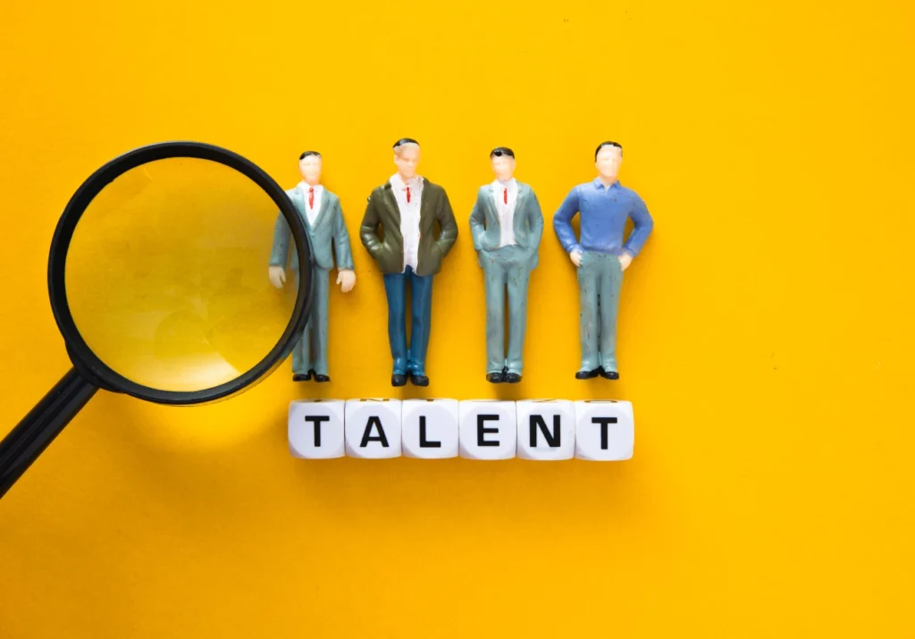 Miniaturas de profissionais alinhadas sobre blocos com a palavra “TALENT”, acompanhadas de uma lupa sobre fundo amarelo, simbolizando identificação e visibilidade de habilidades.
