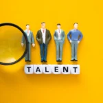 Miniaturas de profissionais alinhadas sobre blocos com a palavra “TALENT”, acompanhadas de uma lupa sobre fundo amarelo, simbolizando identificação e visibilidade de habilidades.
