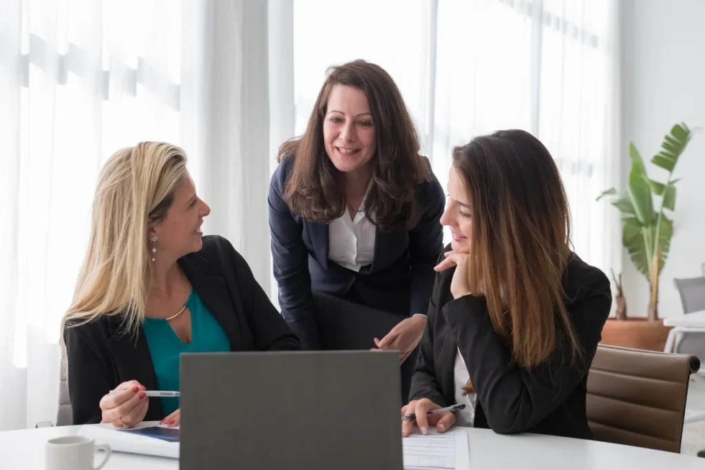 Três profissionais em blazers conversando ao redor de um laptop em sala de reunião clara, com plantas e luz natural ao fundo.