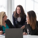 Três profissionais em blazers conversando ao redor de um laptop em sala de reunião clara, com plantas e luz natural ao fundo.
