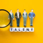 Miniaturas de profissionais alinhadas sobre blocos com a palavra “TALENT”, acompanhadas de uma lupa sobre fundo amarelo, simbolizando identificação e visibilidade de habilidades.