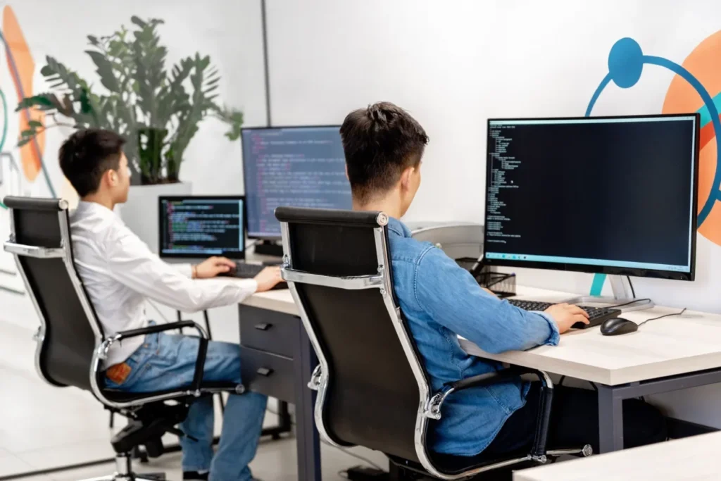 Dos desarrolladores escribiendo código en monitores grandes en una oficina moderna, representando equipos de tecnología y software