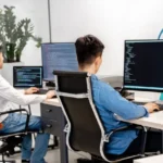 Dos desarrolladores escribiendo código en monitores grandes en una oficina moderna, representando equipos de tecnología y software