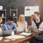 Equipe reunida em uma sala de reunião colaborativa, usando laptops e discutindo estratégias de trabalho em um ambiente corporativo moderno.